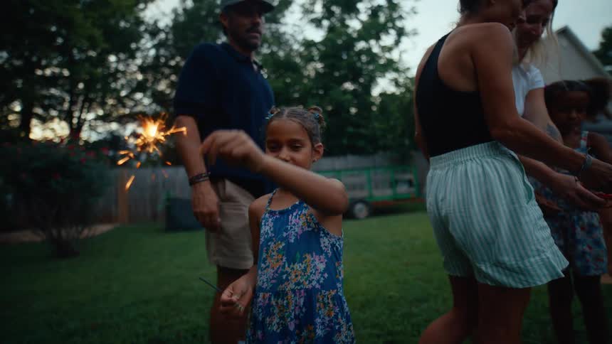A young girl in a floral dress waves a lit sparkler while adults and other children gather around in the yard