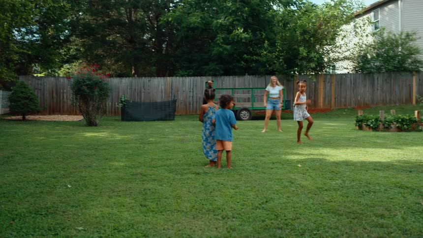 A golden doodle dog and a family of men women and children are playing in a green backyard with a wooden fence and large trees in the background