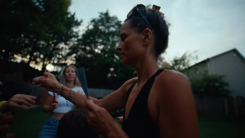 A group of children and adults are gathered in a backyard at dusk a woman in a black tank top is holding a bundle of sparklers and giving one to a young boy