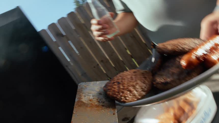 Person barbecuing burgers and sausages and serving it to two adult men standing around holding drinks outdoors during day time