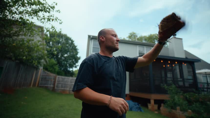 A man in his backyard tosses a baseball to a kid and catches it back in a playful toandfro while others stand around the yard enjoying the sunny midday atmosphere