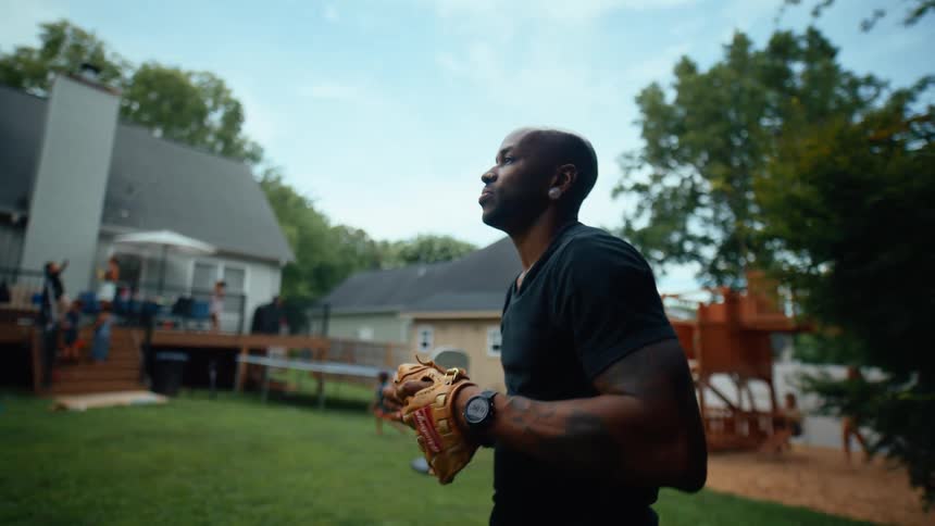 Two men toss a baseball to each other in a backyard at midday while kids run around and other adultsfathers and mothersmingle during the event