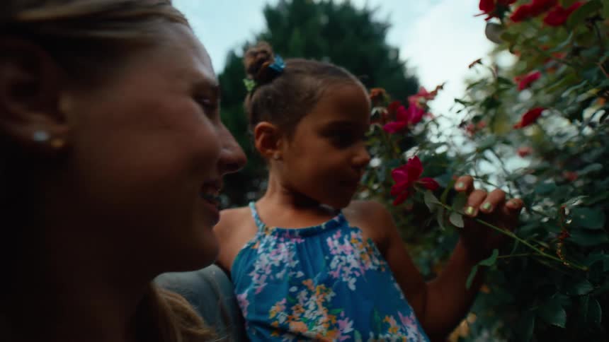 A mother and child in a garden at midday both leaning toward a flower plant smiling with excitement as they breathe in its scent under the bright sun