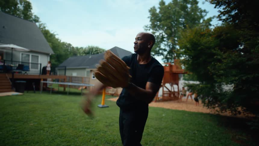A bald man wearing a black shirt and jeans is playing catch with a baseball glove in a suburban backyard with family members and children visible in the background