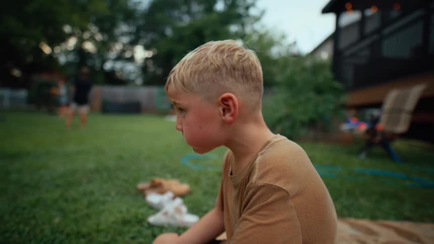A young boy eating a snack in a backyard while other people are playing in the background