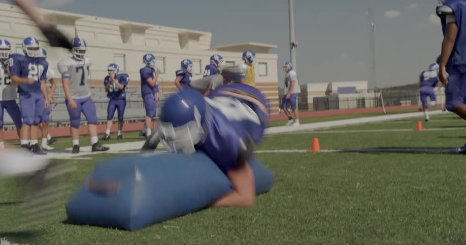 The video shows a football training drill on a field in the foreground a player in a blue jersey with a white number 36 is running toward the left he is about to make contact with a blocking sled in the background a line of players in white jerseys w