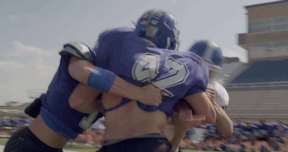 Two football players in blue jerseys and helmets engage in a tackle during practice on a sunny field with other players and spectators in the background