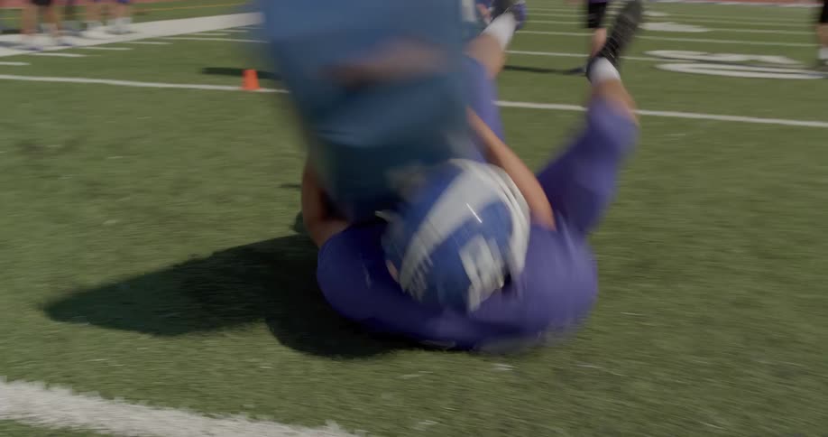The video shows two lines of people at a football practice in the foreground a player in a blue jersey is running and pushing a large padded blue blocking bag in the background a line of other football players in similar blue jerseys stands on the fi
