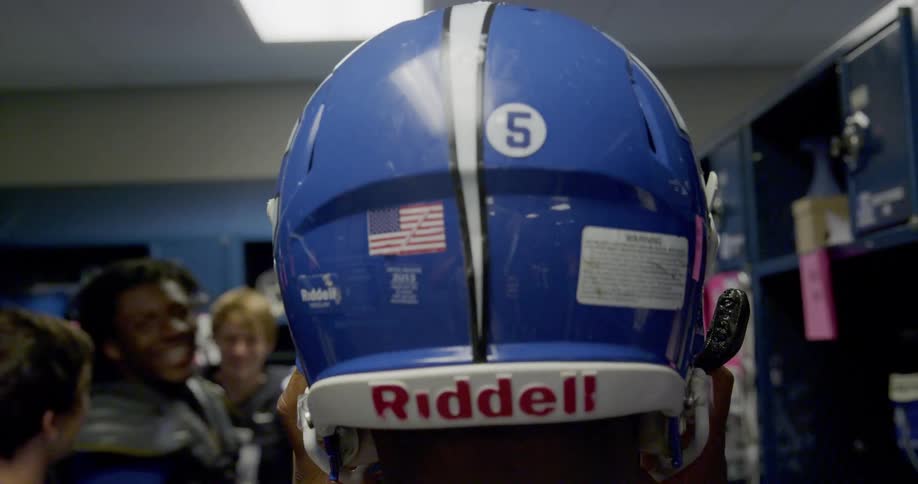 A football player puts on a blue riddell helmet with the number 5 an american flag sticker and a warning label in a locker room with other players visible in the background