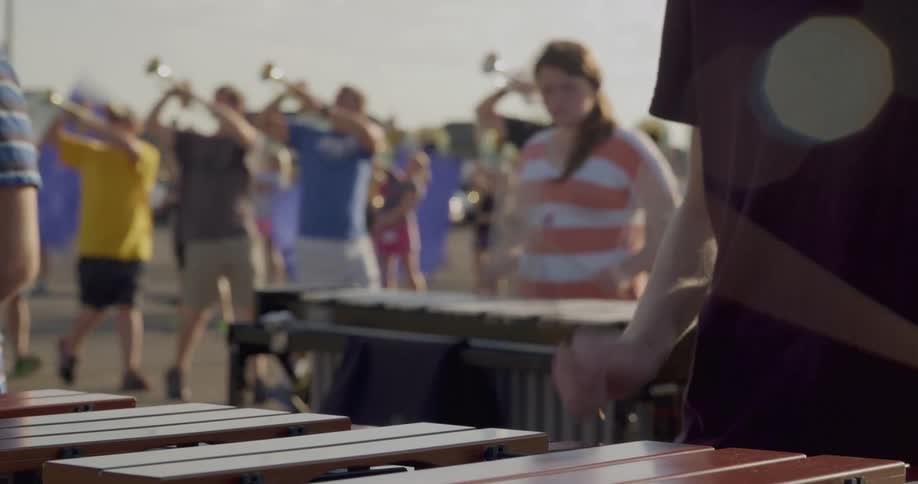 The video shows a musical practice with two lines of people in the foreground a musician is playing a mallet instrument like a marimba with their back to the camera in the background there are two distinct lines of people one line is playing brass