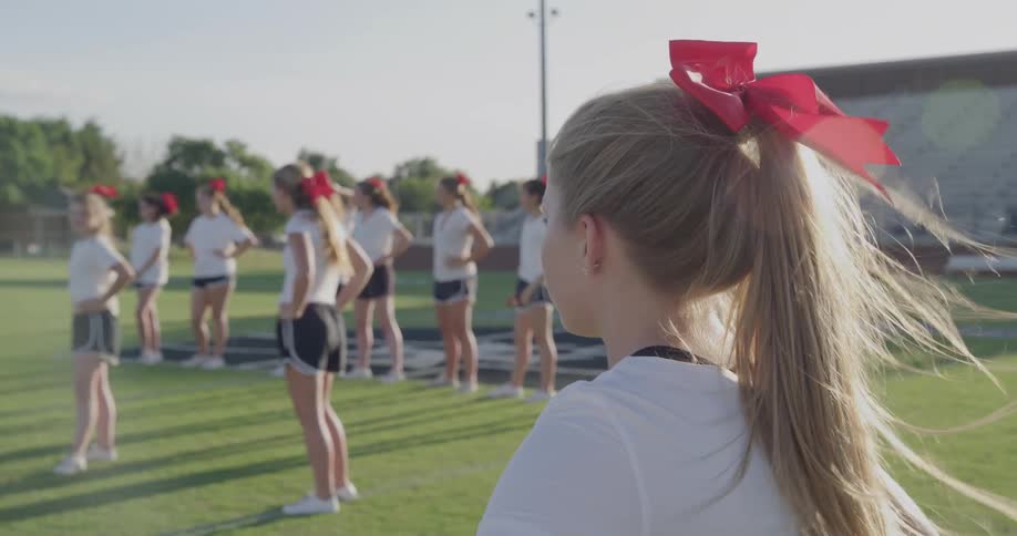 A group of teenage cheerleaders stand on a football pitch on a sunny day wearing black shorts and white tops
