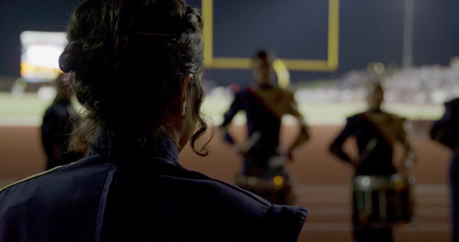 Marching band musicians rehearse on the side of a football field. at night in the foreground a musician with her back to the camera stands observing in the background there is a line of musicians including some drummers wearing black and gold uniforms.