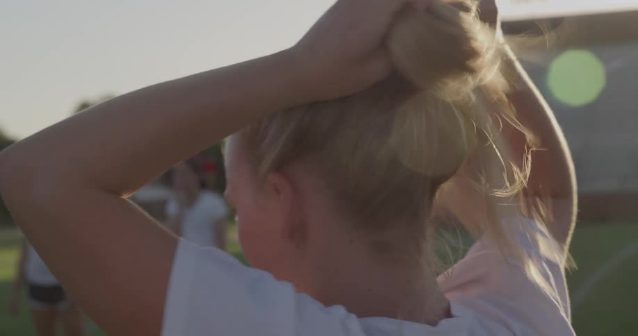 One of the girls standing in shorts on the football field puts her hair into a ponytail
