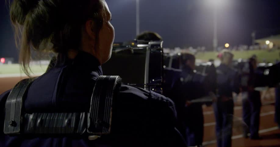 A person in a marching band uniform with a drum strapped to them stands on a track under stadium lights at night with other band members in the background