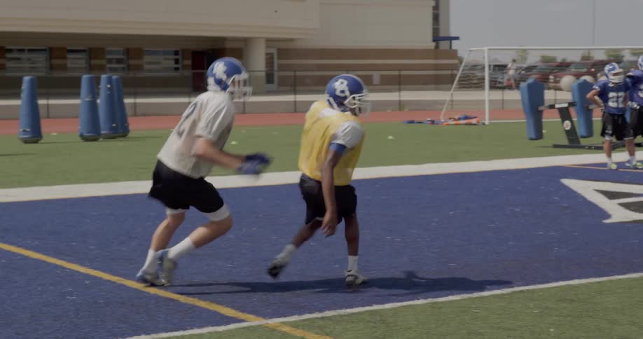 The video shows a football practice with two distinct lines of players in the foreground two playersone in a white jersey and one in a yellow pinnieare running a drill in the background two lines of other players in similar uniforms are facing each