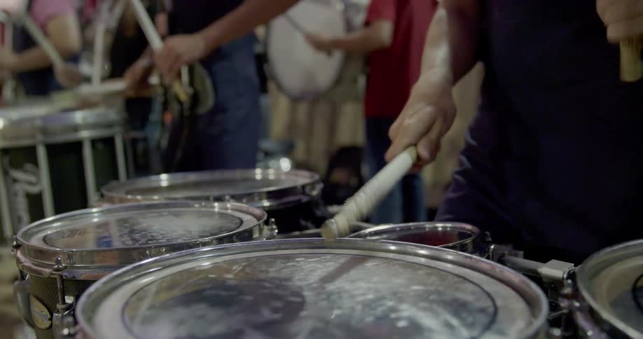 The video shows two lines of musicians with a focus on a drummer in the foreground he is playing a set of drums with drumsticks in the background there is a second line of musicians standing behind the drummer and they appear to be part of a band or