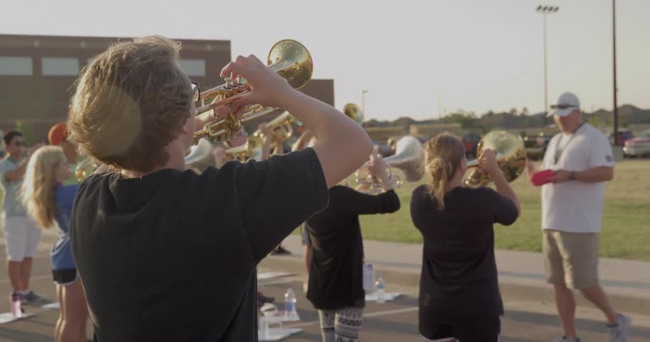 A group of teenagers stand on a football field at midday playing trumpets together during a lesson