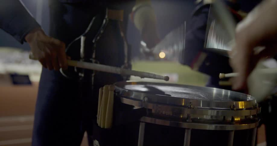 The video shows two lines of musicians with a focus on a drummer in the foreground he is playing a set of drums with drumsticks in the background there is a second line of musicians standing behind the drummer and they appear to be part of a band or