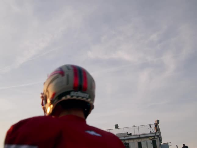 The video shows two distinct lines of football players in uniforms in the foreground a player in a red jersey with a red and gray striped helmet is facing away from the camera in the background a stationary line of players in the same uniform stands o