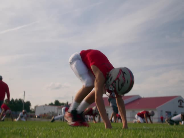 A lowangle eyelevel view shows a high school football player in a red jersey and helmet on a practice on a field with other teammates