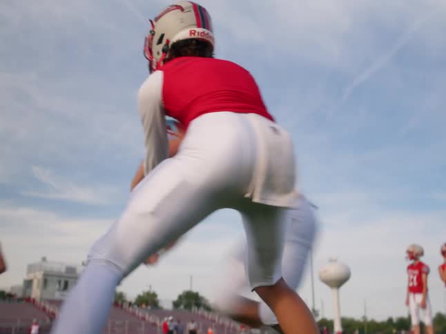 The video shows two lines of people at a football practice in the foreground a football player in a red jersey and white pants is running with a football in the background there is another line of players in similar uniforms