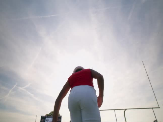 A male high school football player in a red jersey and white pants stands on a green field and stretches his leg with a scoreboard and goalposts in the background under a cloudy evening sky