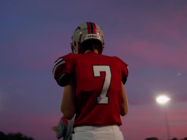 Football player wearing a red jersey with the number 7 on the back and a helmet standing on the field at night during a football game
