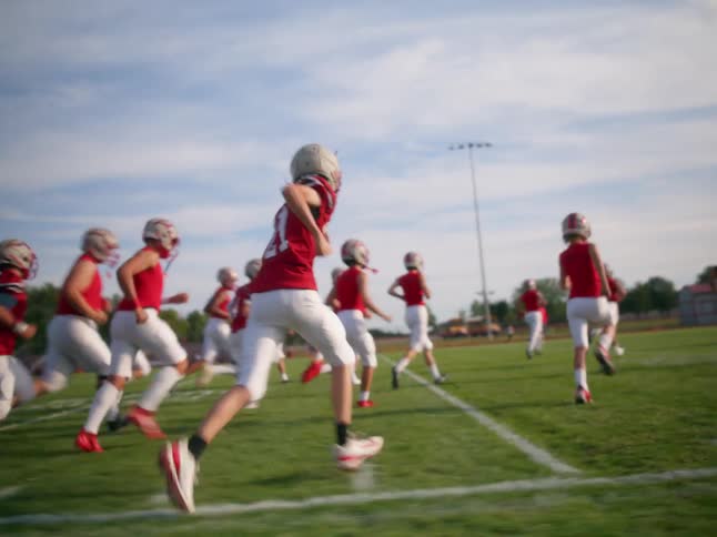 The video shows a football team running a drill on a field in the foreground a line of players in red jerseys and white pants is running toward the right side of the frame in the background a second parallel line of players in the same unifor