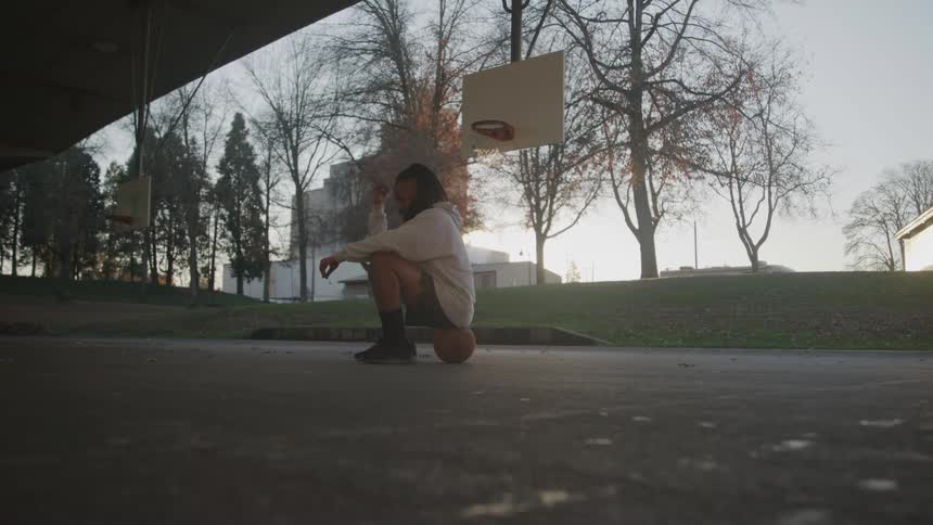 A man with dreadlocks and a long beard is sitting on the ground with a basketball between his legs on a cloudy day with a basketball hoop and trees visible in the background