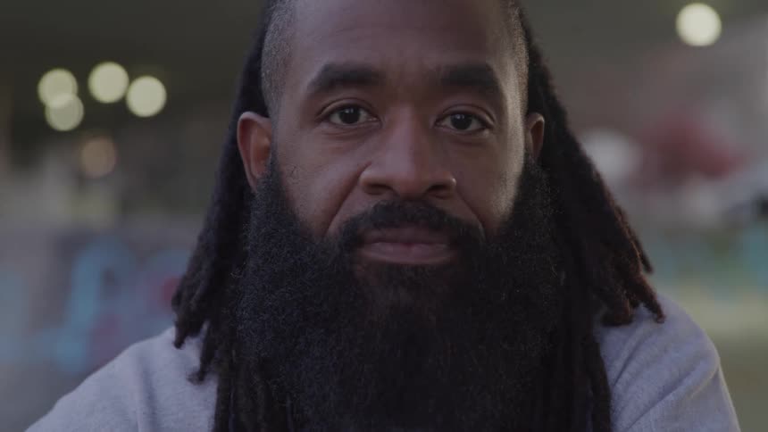 Young black man with long braided thick hair long black beard and mustache standing outdoors at midday under an overhead bridge