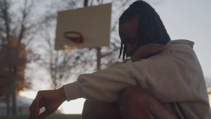 A man with dreadlocks and a beard sits dejectedly on an outdoor basketball court