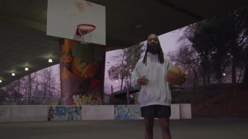 A young black man with long hair in thick braids and a full black beard stands on a street basketball court at evening time casually tossing a basketball from his left hand to his right under the warm glow of the setting sun