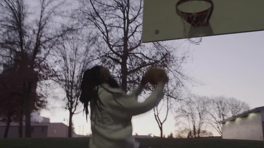 A young person with long hair and a lightcolored sweatshirt dribbles a basketball on an outdoor court they jump up and shoot the ball which goes into the hoop