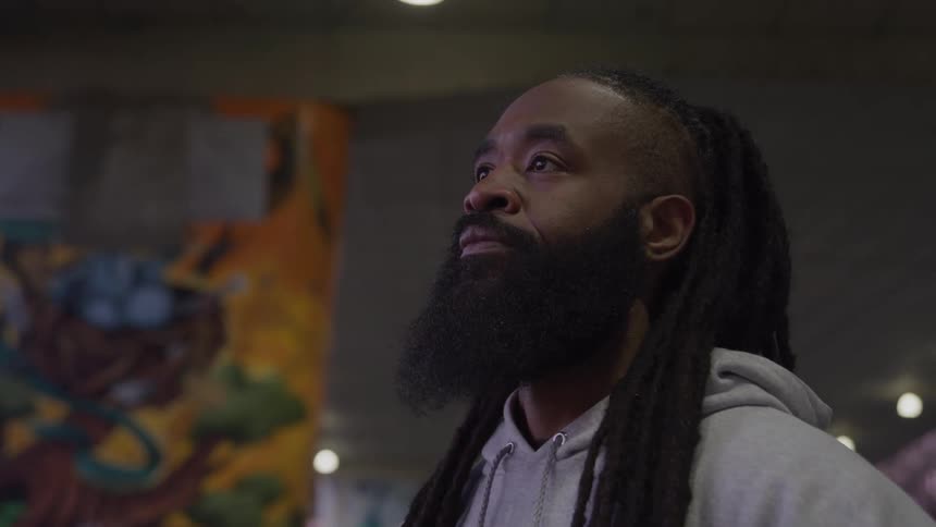A man with a beard and dreadlocks stands holding a basketball looking up and away with colorful graffiti in the background