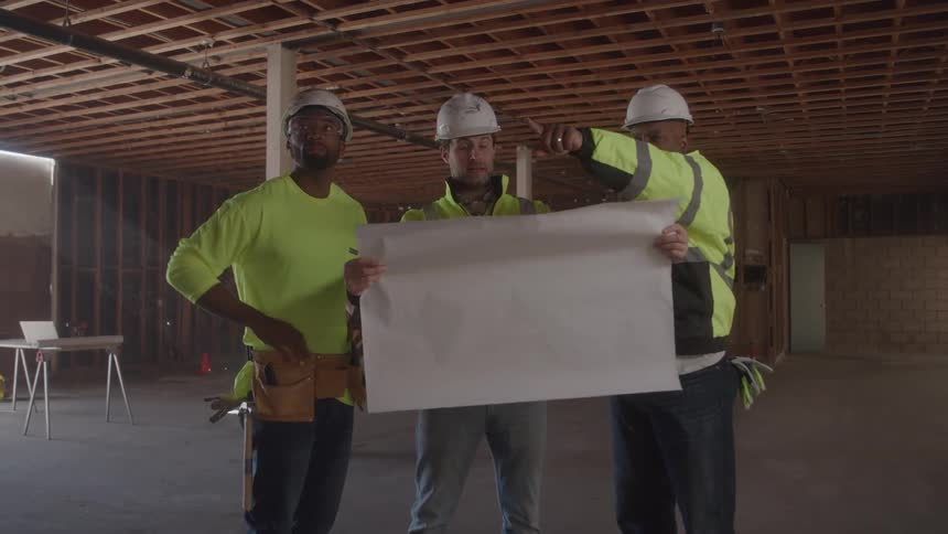 Three male construction workers wearing hard hats and reflective vests stand in an unfinished building and look at blueprints
