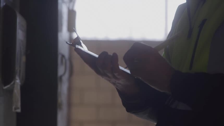 A worker wearing a safety vest and a dark longsleeved shirt holds a clipboard and a pencil the person is focused on what they are writing taking notes or making calculations
