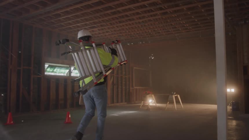 Male construction worker at a framed house carrying a set of stairs on his shoulder indoors during day time
