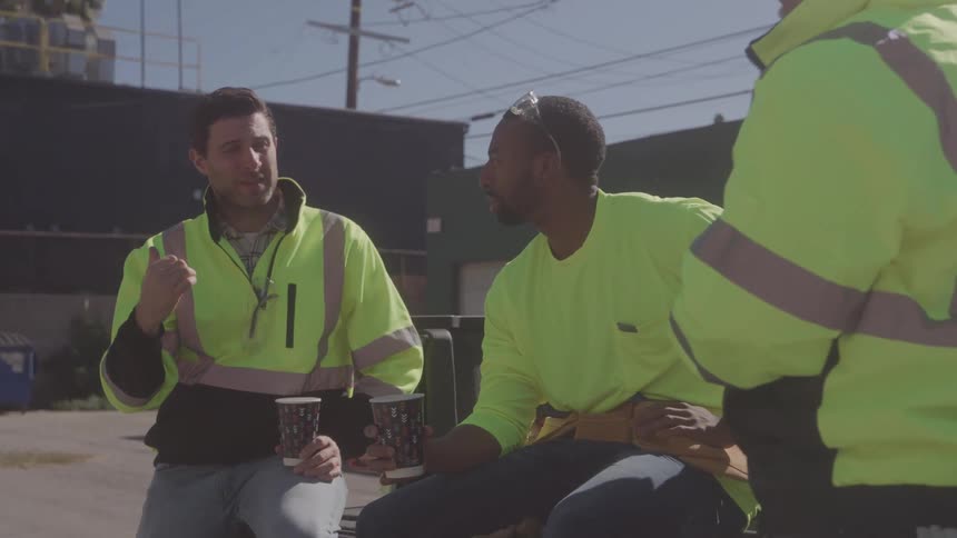 Three workers sit outdoors under the bright sun all wearing safety vests and holding cups of coffee engaged in conversation