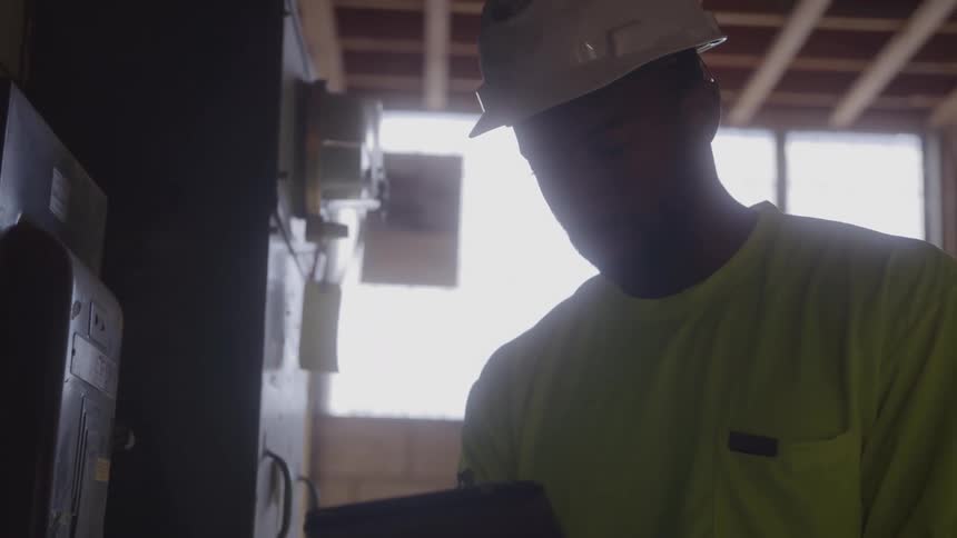 A man wearing a hard hat and a bright yellow shirt stands in front of a window and looks at a device or a clipboard