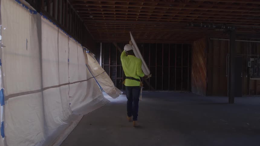 Construction worker wearing a hard hat and green safety vest carrying a plain board while walking indoors at a construction site