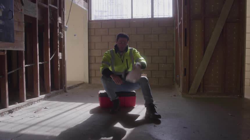 A tired construction worker in a hard hat and reflective vest sits on a cooler in an empty partially demolished building removing his hat and wiping his forehead in the bright sunlight