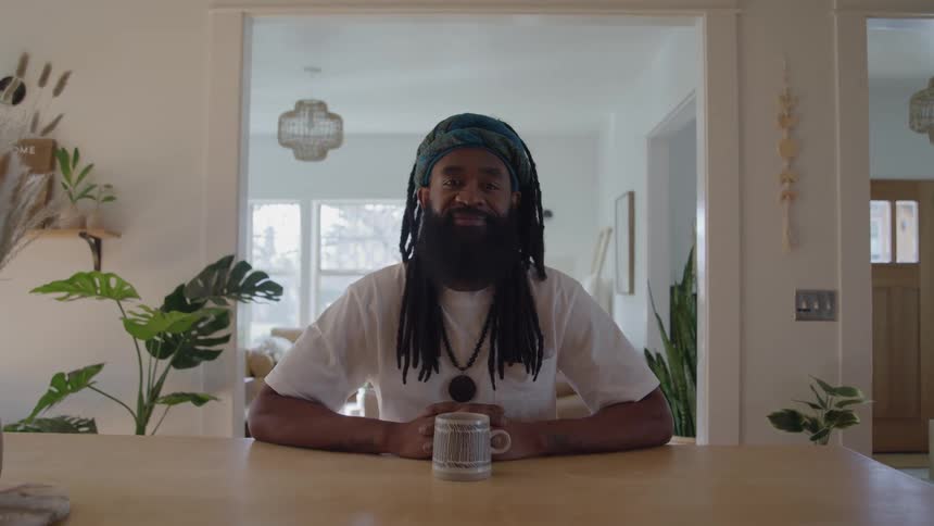 A bearded man with dreadlocks and a blue head wrap is sitting at a wooden table holding a cup and looking directly at the camera in a modern home with potted plants and sunlight coming through a window in the background