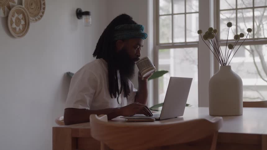 A man with a beard and dreadlocks is working on a laptop at a wooden table in a brightly lit room taking a moment to drink from a cup