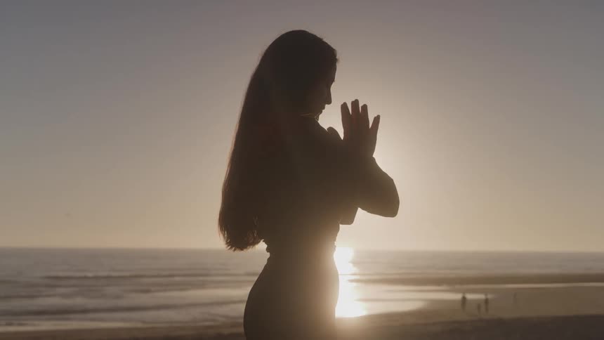 A silhouette of a woman with long hair stands on a beach with her hands in a prayer pose with the sun setting over the ocean behind her