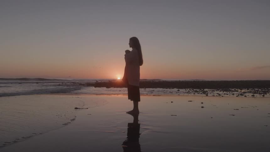 A silhouette of a barefoot woman stands on a wet sandy beach at sunset with her hands together near her chin and her hair blowing slightly in the wind