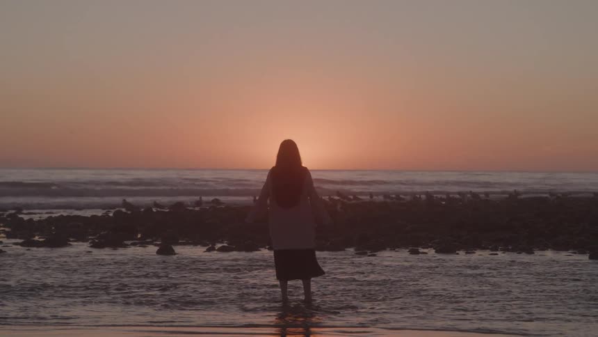 Woman walking in shallow water on the beach with arms stretched out facing the orange setting sun in the distance