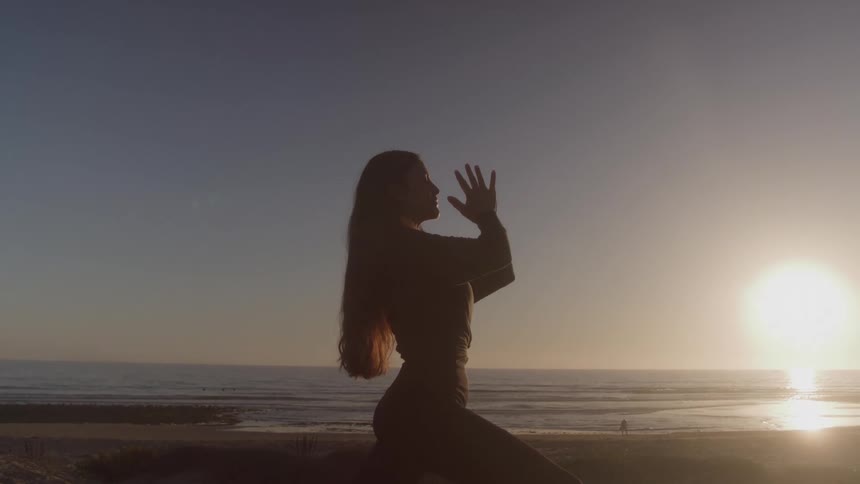 A female yoga enthusiast with long hair is doing a yoga pose on a beach at sunset with her hands raised and her body silhouetted against the bright light and ocean