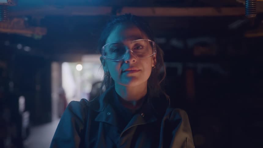 Woman in overalls and safety glasses looks at the camera inside the workshop