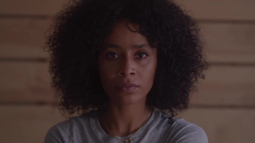 A medium closeup shot of a woman with curly brown hair looking directly at the camera she has a neutral expression on her face and is standing in front of a light wooden wall