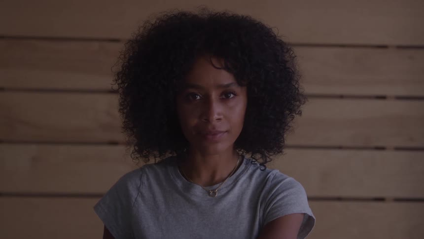 A medium closeup shot of a woman with curly brown hair standing in front of a light wooden wall she turns her head to look directly at the camera and smiles slightly